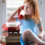 Small Talk Conversation - Low angle of calm redhead preteen lady in blue dress and beige sandals looking away and having phone call using retro disk telephone on stack of books while sitting with legs crossed on wooden table against window at home