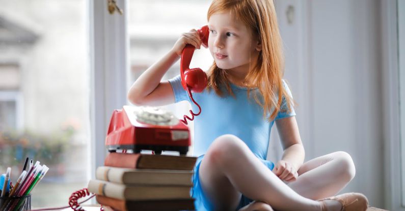 Small Talk Conversation - Low angle of calm redhead preteen lady in blue dress and beige sandals looking away and having phone call using retro disk telephone on stack of books while sitting with legs crossed on wooden table against window at home