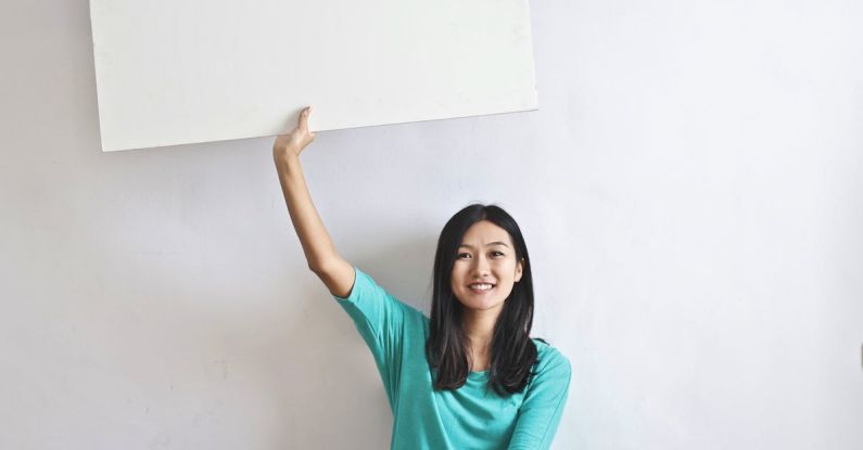Influencer Marketing - Cheerful Asian woman sitting cross legged on floor against white wall in empty apartment and showing white blank banner