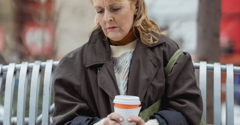 Upselling Cross-selling - Lonely elderly female in coat with hot drink to go sitting with crossed legs on bench while looking down