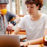 Remote Teams - Pensive young woman typing on netbook while man with cup drinking coffee at table of cafe